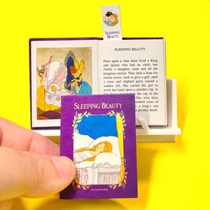 Peut inclure: Un livre miniature intitulé "Sleeping Beauty" avec une couverture violette, tenu devant un livre ouvert. L'illustration de la couverture représente une femme endormie. Le livre ouvert affiche du texte et des illustrations. Un marque-page avec le titre est également visible.