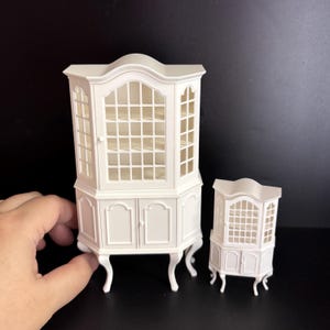 Puede incluir: Dos armarios de esquina en miniatura blancos. El armario más grande tiene una sección superior con frente de vidrio con múltiples estantes y una sección inferior con dos puertas. El armario más pequeño refleja el diseño. Ambos tienen patas curvas.