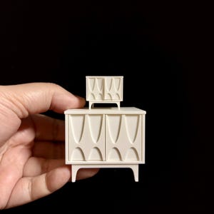 Puede incluir: Un juego de muebles en miniatura de estilo moderno de mediados de siglo, de color crema. El mueble más grande tiene dos puertas con un diseño curvo, y el mueble más pequeño está encima. Ambos tienen patas cortas y anguladas.