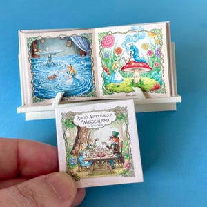 May include: Miniature book set of Alice's Adventures in Wonderland. The set includes a small book held by a hand, and a larger open book with illustrations of Alice in Wonderland scenes. The cover of the small book reads "Alice's Adventures in Wonderland" by Lewis Carroll.