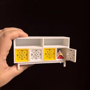 May include: A miniature white cabinet with yellow and white decorative door panels. One door is open, revealing a small figure in traditional Korean attire. The cabinet is held by a hand against a dark background.