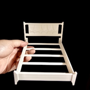 May include: A small, off-white bed frame with a decorative headboard. The headboard features a series of arched designs. The bed frame has a rectangular shape with four horizontal support beams. The bed frame is held by a hand against a black background.