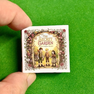 May include: A miniature book titled "The Secret Garden" by Frances Hodgson Burnett. The cover features an illustration of three children holding hands, surrounded by floral archways. The book is held in a person's hand against a green background.