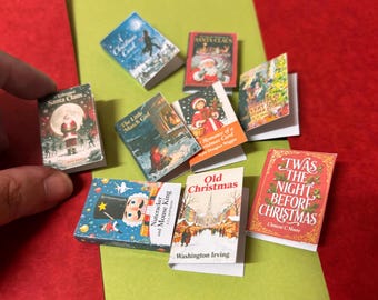 May include: A collection of miniature Christmas-themed books with colorful covers. Titles include "Santa Claus," "Christmas Carol," "The Little Match Girl," and "Twas the Night Before Christmas." The books are arranged on a red and green surface.