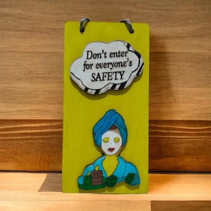 Puede incluir: Un letrero de madera amarillo con una nube blanca y texto negro que dice "No entres por la seguridad de todos". Debajo de la nube hay una ilustración de dibujos animados de una persona con un turbante azul y una máscara facial blanca.