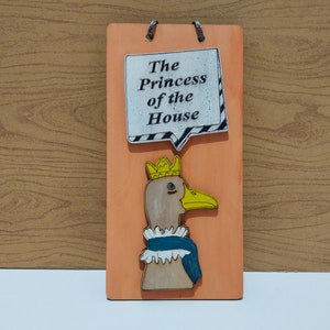 May include: A wooden sign with a peach background and a cartoon duck wearing a crown and a blue scarf. The sign says "The Princess of the House" in a speech bubble.