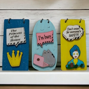 Könnte beinhalten: Drei dekorative Holzschilder mit humorvollen Botschaften. Eines ist blau mit einer Krone und dem Text "The Princess of the House". Ein weiteres ist hellblau mit einer Katze und dem Text "I'm busy". Das dritte ist gelb mit einer Person und dem Text "Don't enter for everyone's SAFETY."