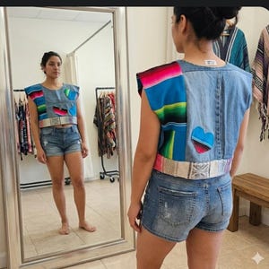 May include: A denim crop top with colorful woven fabric accents and a heart-shaped patch. The top features a woven belt and is paired with denim shorts. The top has a unique, patchwork design.