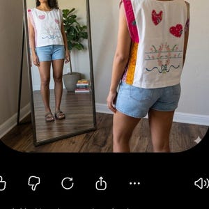 May include: A white sleeveless top featuring colorful patchwork side panels and embroidered heart and floral motifs on the back. The garment is paired with light blue denim shorts. The design includes decorative stitching and fabric accents.