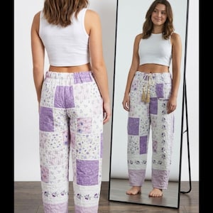 May include: A white sleeveless crop top and quilted pants featuring a patchwork design in shades of purple, lavender, and white with floral patterns. The pants have an elastic waistband with a drawstring featuring two hanging tassels.