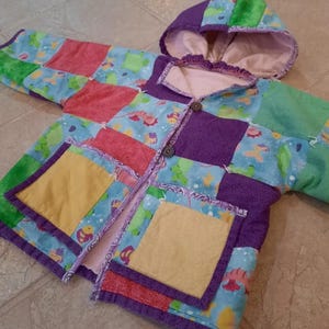 May include: A quilted jacket for a child with a hood, featuring a patchwork design of various colors, including purple, green, blue, yellow, and pink. The jacket has two pockets and a button closure.