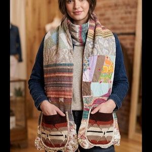 May include: A long scarf featuring a patchwork design of various fabric patterns including floral, plaid, and geometric prints. The scarf has a high neck collar and two large, dark brown and cream-colored crocheted pockets with a geometric pattern.