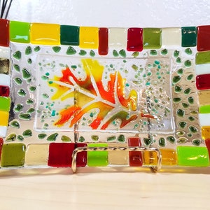 May include: A rectangular glass dish with a mosaic border of red, green, yellow, and white tiles. The center of the dish features a painted image of an orange, yellow, and green leaf.