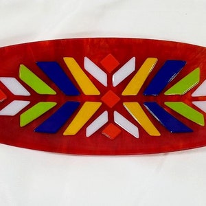 May include: An oval-shaped, red glass dish with a geometric design in the center. The design features white, yellow, green, blue, and red pieces of glass.