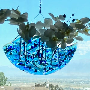May include: A hanging glass art piece with a blue and white design, holding white orchid flowers. The glass has a semi-circular shape with blue and dark blue speckles. The piece hangs in front of a window.