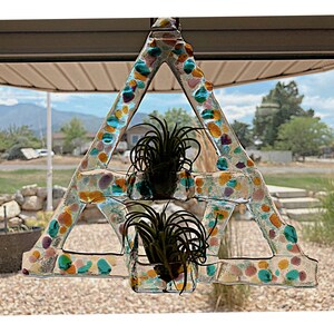 May include: A glass triangle-shaped planter with colorful flecks of glass. Two air plants are growing in the planter. The planter is hanging from a window.