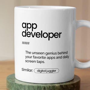 May include: White ceramic mug with the text "app developer" and a definition. The mug sits on a green coaster atop a wooden table. The text on the mug reads "The unseen genius behind your favourite apps and daily screen taps."