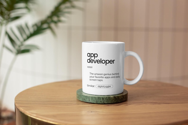 May include: White ceramic mug with the text "app developer" and a definition. The mug sits on a green coaster atop a wooden table. The text on the mug reads "The unseen genius behind your favourite apps and daily screen taps."