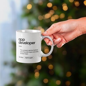May include: White ceramic mug with the text "app developer" and a definition. The mug is held by a hand against a blurred background of green and gold lights. The definition reads, "The unseen genius behind your favourite apps and daily screen taps."