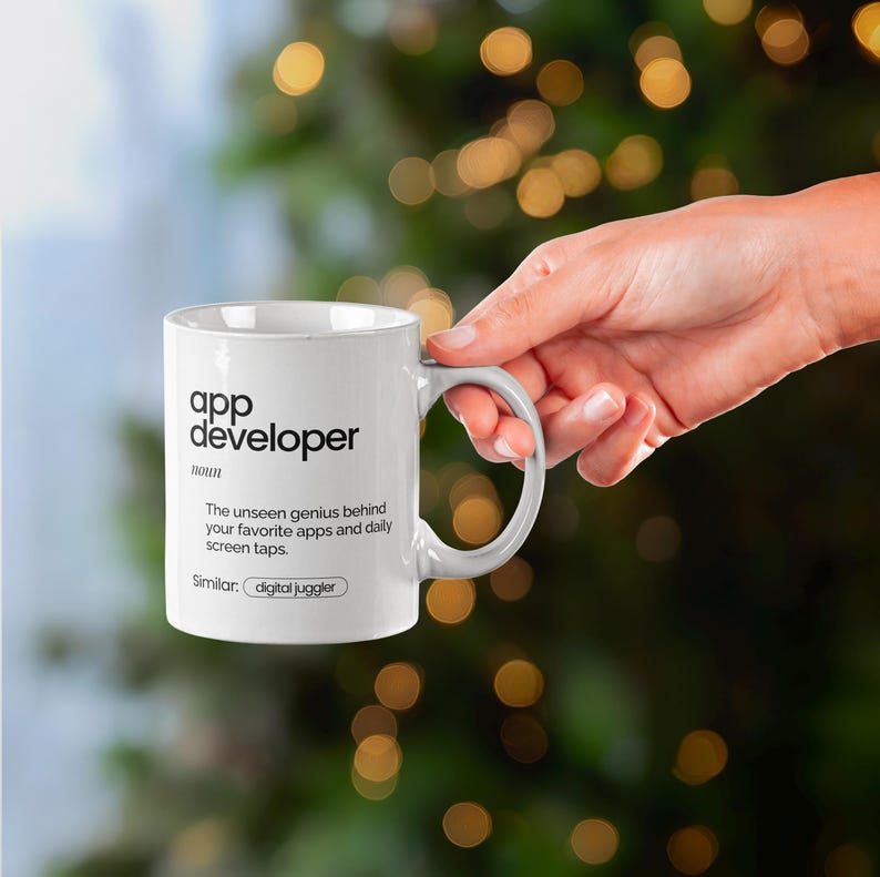 May include: White ceramic mug with the text "app developer" and a definition. The mug is held by a hand against a blurred background of green and gold lights. The definition reads, "The unseen genius behind your favourite apps and daily screen taps."
