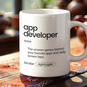 May include: White ceramic mug with the text "app developer" in black, and a definition. The mug also includes the phrase "The unseen genius behind your favourite apps and daily screen taps." and the word "Similar: digital juggler."