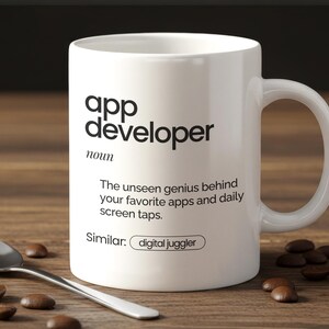 May include: White ceramic mug with the text "app developer" in black. The mug also has the definition of app developer printed on it. A silver spoon and coffee beans are on the wooden surface.