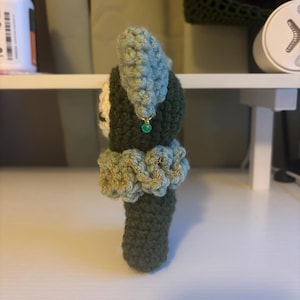 Puede incluir: Mu&ntilde;eca de ganchillo hecha a mano en tonos verdes, azules y crema. La mu&ntilde;eca tiene un cuello con volantes con detalles dorados y una peque&ntilde;a cuenta verde. El sombrero de la mu&ntilde;eca es de color azul claro.