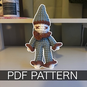 Peut inclure: Poupée crochetée avec un chapeau pointu, une tenue bleu-gris et des accents marron. La poupée a un visage blanc avec des détails cousus et une écharpe marron. Le texte "PDF PATTERN" est affiché en bas.