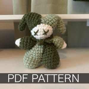 May include: A crochet plush toy featuring a jester-style hat with two long points. The toy has a white face with dark green stitched X-shaped eyes, and a body made of alternating dark green and light green yarn. The text PDF PATTERN is visible.