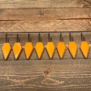 May include: Eight vintage orange-yellow knitting needle holders with metal needles. The holders are diamond-shaped and arranged in a row on a wooden surface. The needles are thin and silver-colored.