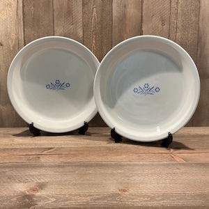 May include: Two round white ceramic plates, each featuring a centered blue floral pattern consisting of small blossoms and leaves. The plates have a smooth, glossy finish and are displayed upright on small black stands, showcasing a simple design.