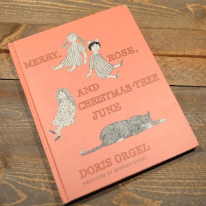 May include: A pink book cover with the title "Merry, Rose, and Christmas-Tree June" by Doris Orgel. The cover features illustrations of three girls and a cat.