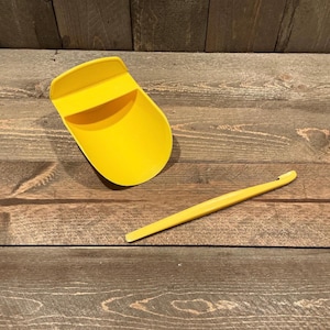 May include: A bright yellow plastic scoop with a wide, open bowl and a flat handle, accompanied by a long, slender yellow plastic tool featuring a small, curved hook at one end, both designed for handling granular materials or small items.