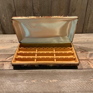 May include: A rectangular jewelry display case with a hinged lid. The interior features a cream-colored satin lining on the lid and a tan, molded velvet-like insert with three horizontal rows of scalloped slots designed for organizing rings.