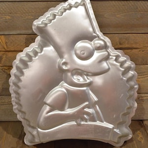 May include: A silver-colored metal cake pan in the shape of Bart Simpson from The Simpsons. The pan features a detailed relief of Bart's face and torso, with a scalloped edge. The pan has a hole at the top for hanging.