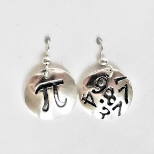 Może przedstawiać: Para srebrnych kolczyków z okrągłym dyskiem. Jeden kolczyk ma wygrawerowany symbol matematyczny pi, a drugi kolczyk ma wygrawerowane pierwsze cyfry pi.