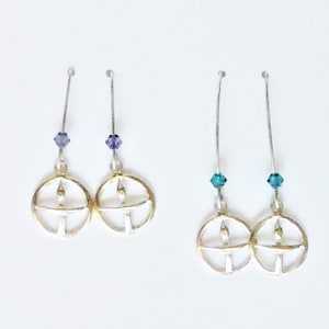 May include: Two pairs of dangle earrings with silver hooks and gold circle charms. One pair has purple crystal accents and the other pair has teal crystal accents.