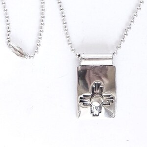 May include: A silver chain necklace with a rectangular pendant featuring a stylized silver  symbol of the Zia sun symbol.