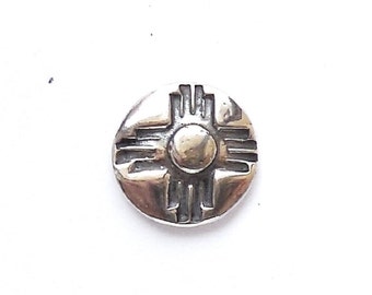 May include: A silver charm with a raised design of a stylized sun symbol, often associated with the Southwest region of the United States.