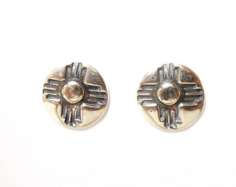 May include: A pair of silver earrings with a gold-toned design of a stylized sun symbol. The earrings are round and have a raised design.