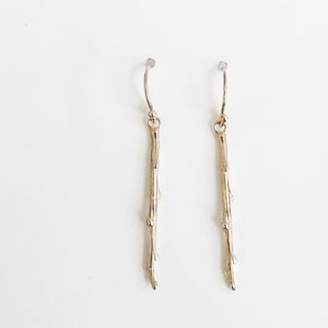 Puede incluir: Un par de pendientes de color dorado en forma de ramitas delicadas con pequeñas ramas. Los pendientes tienen un diseño simple de aro en la parte superior.