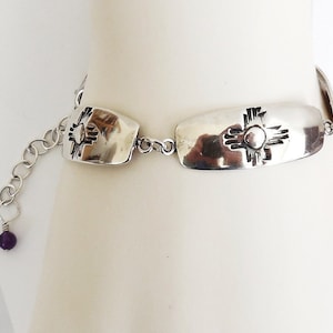 May include: A silver-toned bracelet featuring rectangular links with a sun symbol design. The bracelet has an adjustable chain and a small purple bead at the end. The bracelet is displayed on a white background.