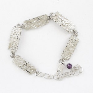 May include: A silver bracelet with a chain and a purple gemstone. The bracelet has five rectangular links with an intricate, textured design.