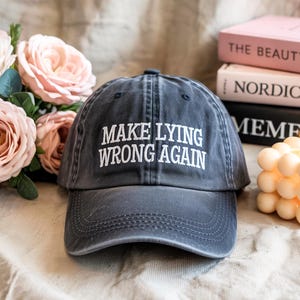 May include: A faded navy blue baseball cap with the text "MAKE LYING WRONG AGAIN" in white. The hat is sitting on a light-colored surface, with pink roses and books in the background.