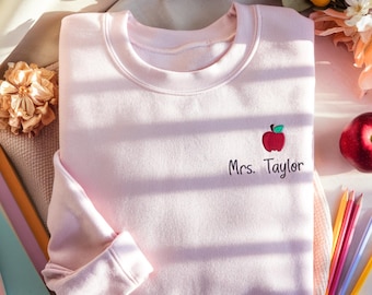 May include: A light pink sweatshirt with a crew neck, featuring a red apple graphic with a green leaf and the name "Mrs. Taylor" embroidered below. A red apple and colored pencils are next to the sweatshirt. The sweatshirt is folded on a soft surface.