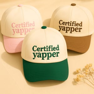 May include: Three baseball caps with the text "Certified yapper". The caps have different color combinations: cream with pink brim, cream with brown brim, and cream with green brim. Each cap has a matching button on top.
