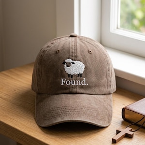 Könnte beinhalten: Eine braune Baseballkappe mit Used-Look, die ein gesticktes weißes Schaf mit schwarzem Gesicht und schwarzen Beinen zeigt. Unter dem Schaf ist das Wort Found. in weißem Garn eingestickt. Die Kappe hat einen gebogenen Schirm und eine feste Krone.