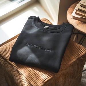 May include: A black crewneck sweatshirt with the embroidered phrase "do it for the plot." The sweatshirt is folded neatly on a corduroy ottoman. The image is lit by natural light.