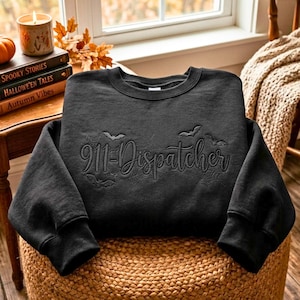 Könnte beinhalten: Ein schwarzes Sweatshirt mit Rundhalsausschnitt und einer tonalen schwarzen Stickerei, die 911-Dispatcher in Schreibschrift zeigt. Das Design umfasst gestickte Spinnweben und kleine fliegende Fledermäuse um den Text für eine dunkle Optik.