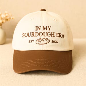 May include: A baseball cap with a cream-colored crown and a brown brim. The cap features the embroidered text "IN MY SOURDOUGH ERA" above an illustration of a loaf of bread, with "EST 2025" below. The top button is brown.